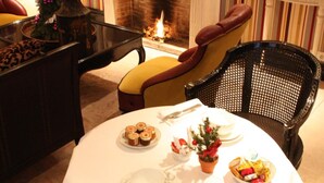 Un comedor con una mesa redonda dispuesta para dos personas, rodeada de sillas de mimbre. Se ve una chimenea al fondo, y las paredes están cubiertas de un papel pintado a rayas.