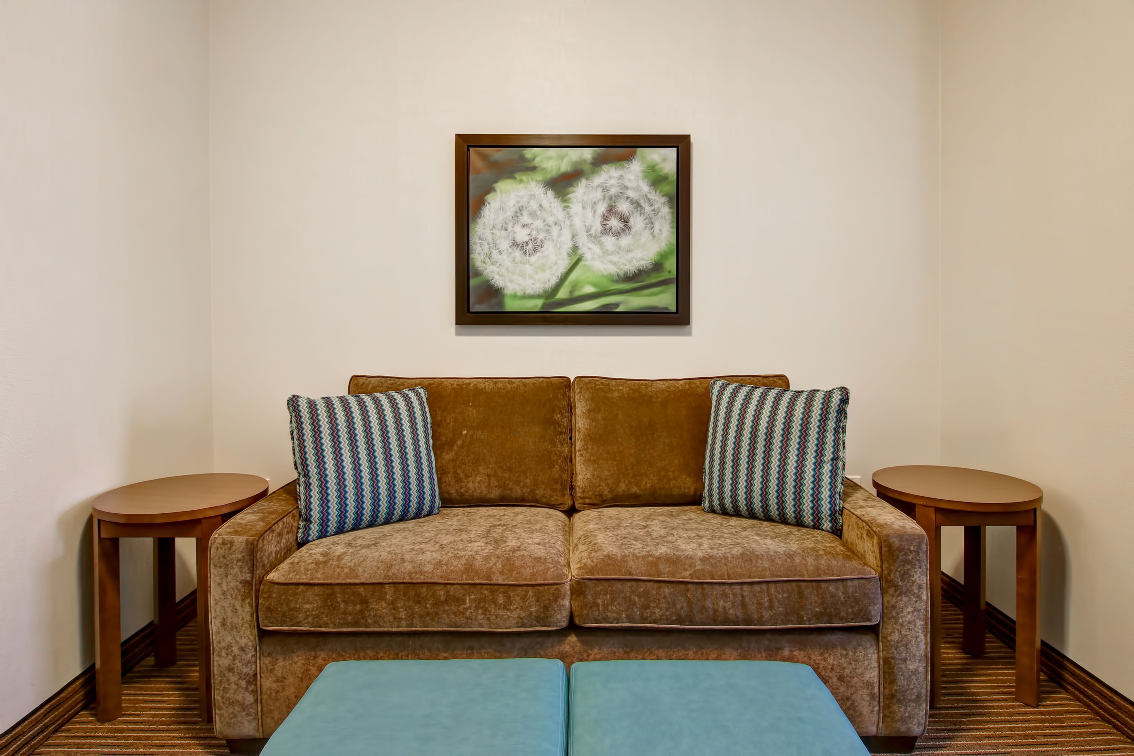 A brown sofa with two patterned pillows, a blue ottoman, and two wooden side tables. A framed picture of dandelions hangs on the wall.