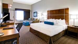 A hotel room with a large bed, a desk with a computer, a chair, a sofa, and a bedside table with a lamp.