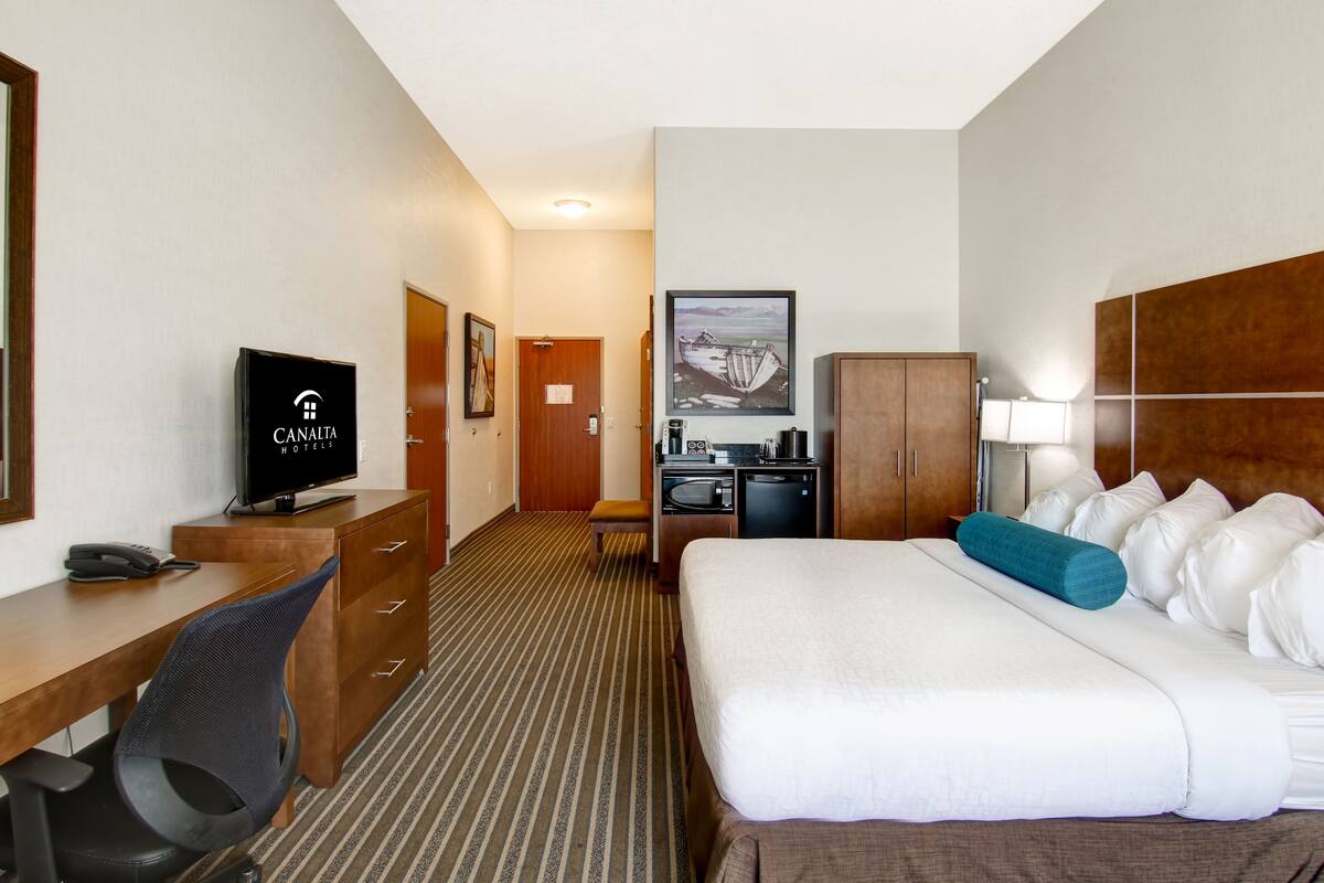 A hotel room with a bed, desk, chair, television, and a kitchenette.