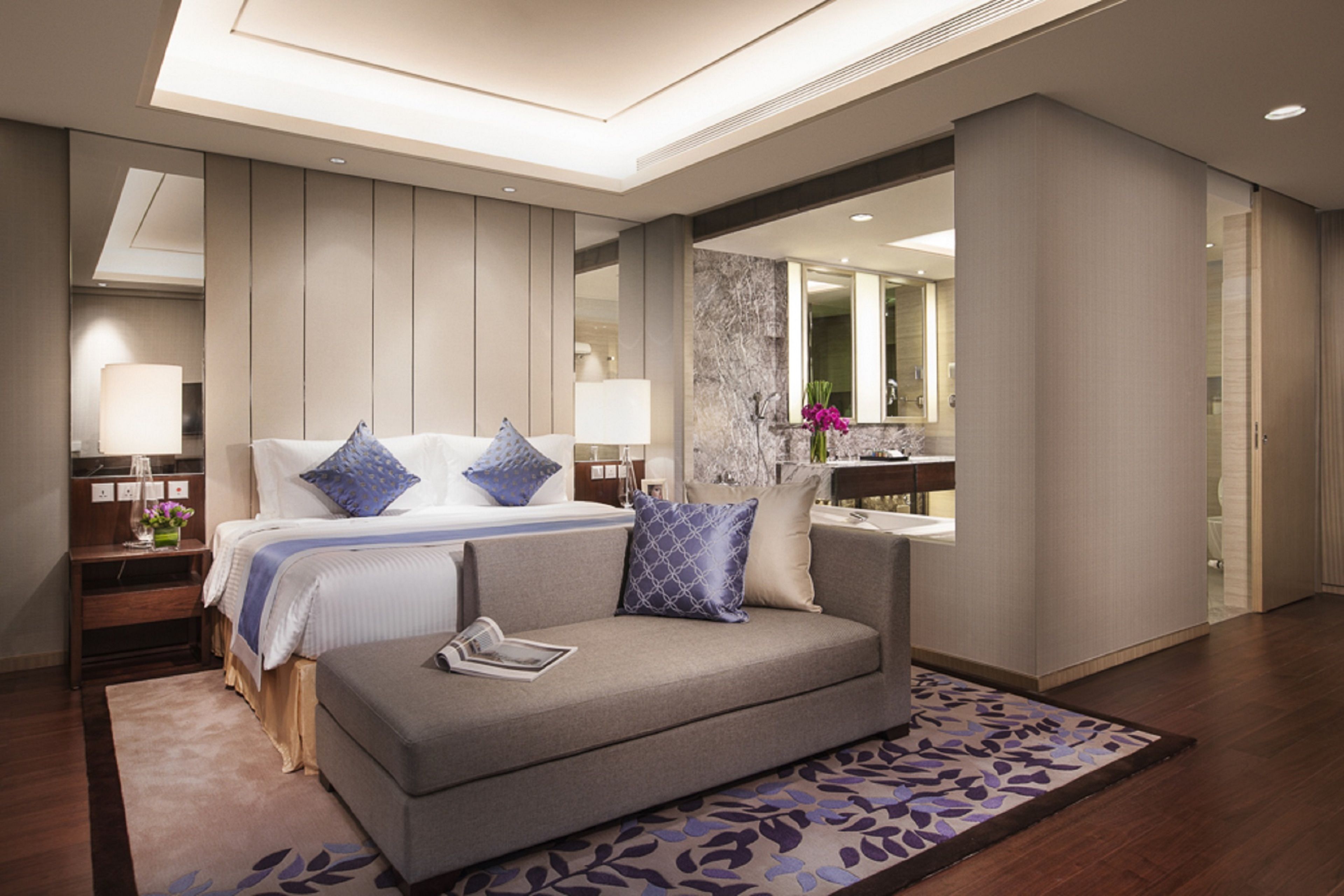 Ein modernes Hotelzimmer mit Bett, Sofa, Nachttisch, Spiegel und einem durchgegangene Tür sichtbaren Badezimmer.