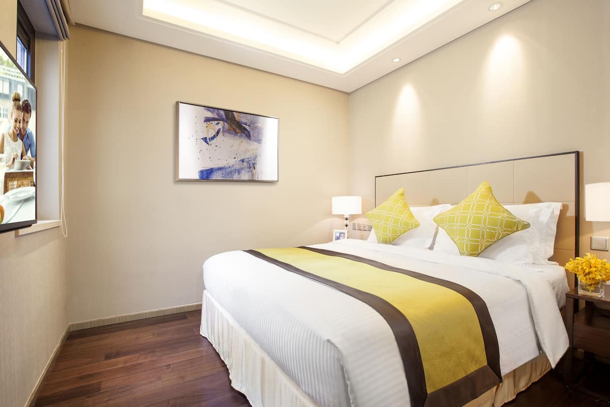 A hotel room with a large bed, a bedside table with a lamp, a wall-mounted painting, and a window with a view of the outside.