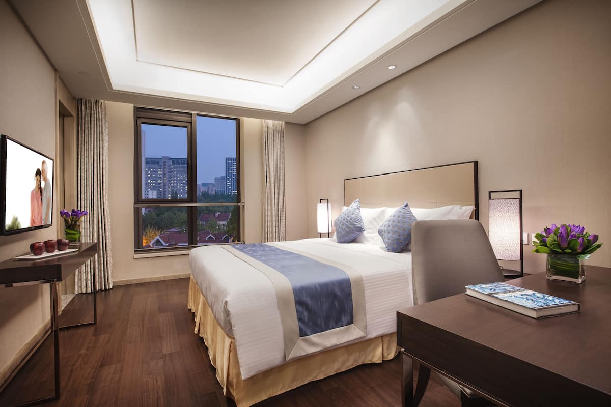 A hotel room with a large bed, a desk, a chair, a TV, and a window with a city view.
