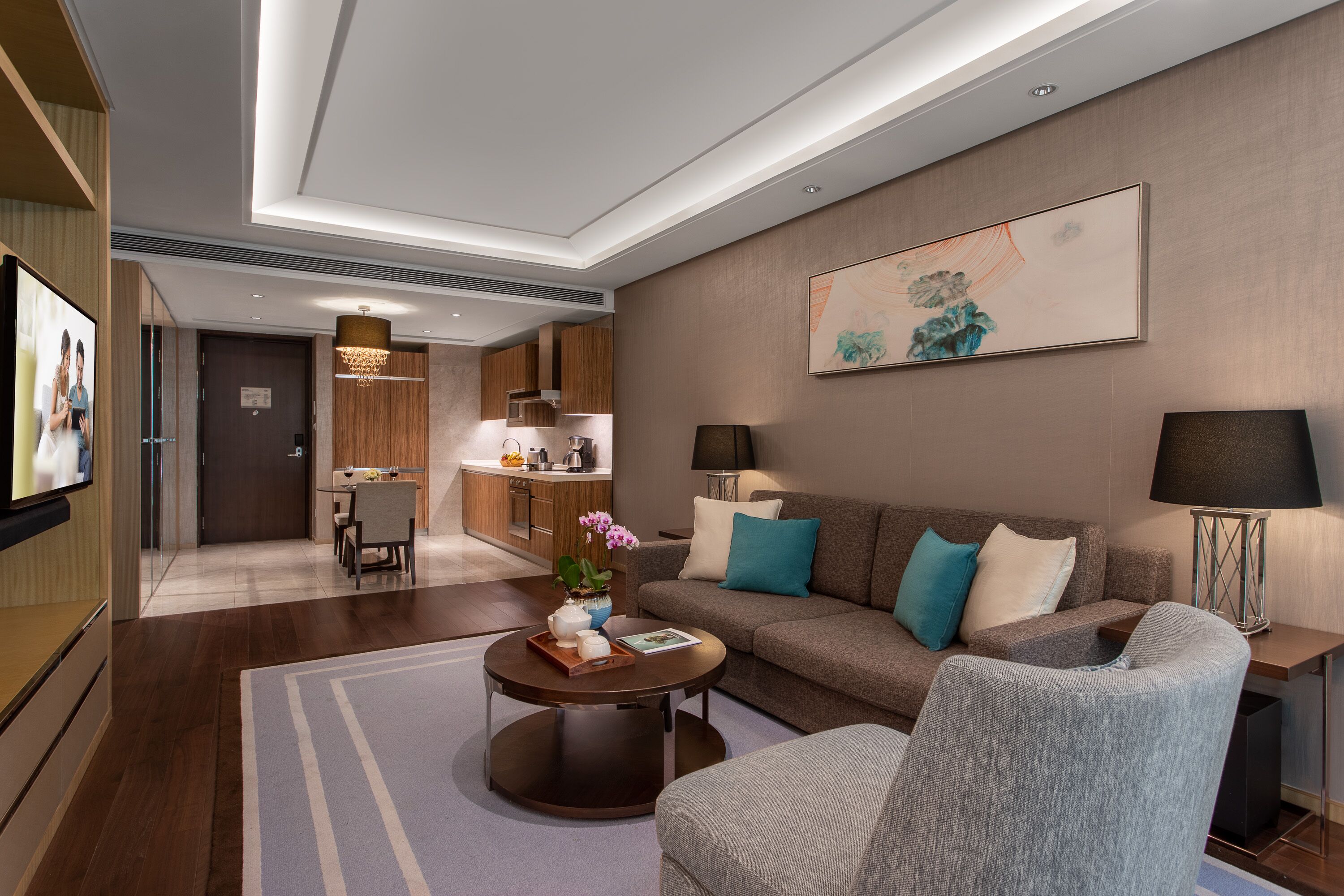 Un salon moderne avec un canapé, des fauteuils, une table basse et un téléviseur. On aperçoit à l’arrière-plan un coin cuisine avec des meubles en bois et une table à manger.