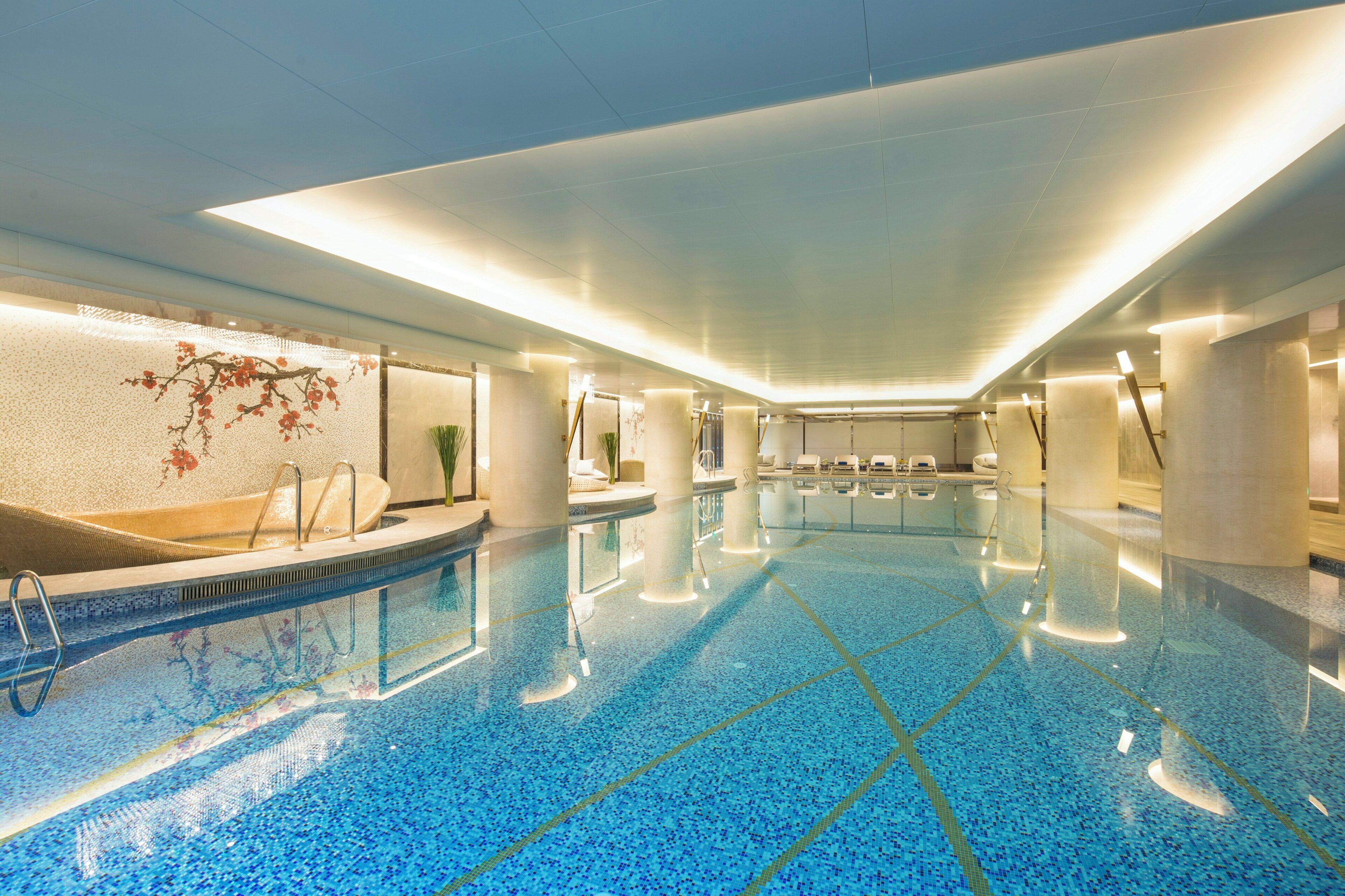 A large indoor swimming pool with clear blue tiles, surrounded by lounge chairs and a wall with a floral design.