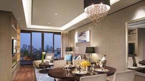 Quarto de hotel moderno com área de jantar, uma grande janela com vista para a cidade, um candelabro e uma televisão de tela plana na parede.
