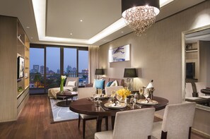 Una habitación de hotel moderna con zona de comedor, un amplio ventanal con vistas a la ciudad, una araña y un televisor de pantalla plana montado en la pared.