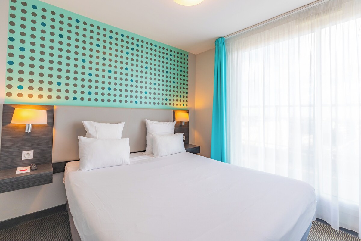 Une chambre d’hôtel avec un grand lit, deux tables de chevet avec des lampes, un mur orné d’un motif à pois et une fenêtre avec des rideaux légers.