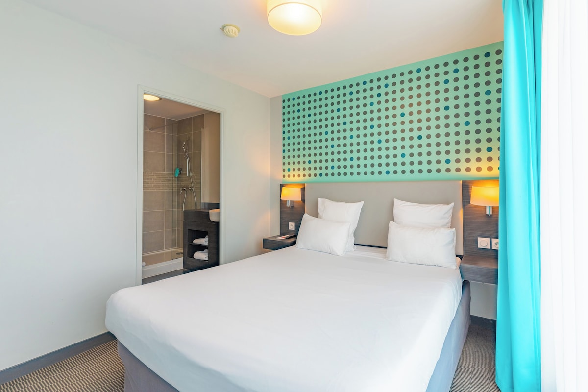 Une chambre d’hôtel avec un grand lit, une douche, un luminaire fixé au mur et une tête de lit ornée d’un motif à pois.