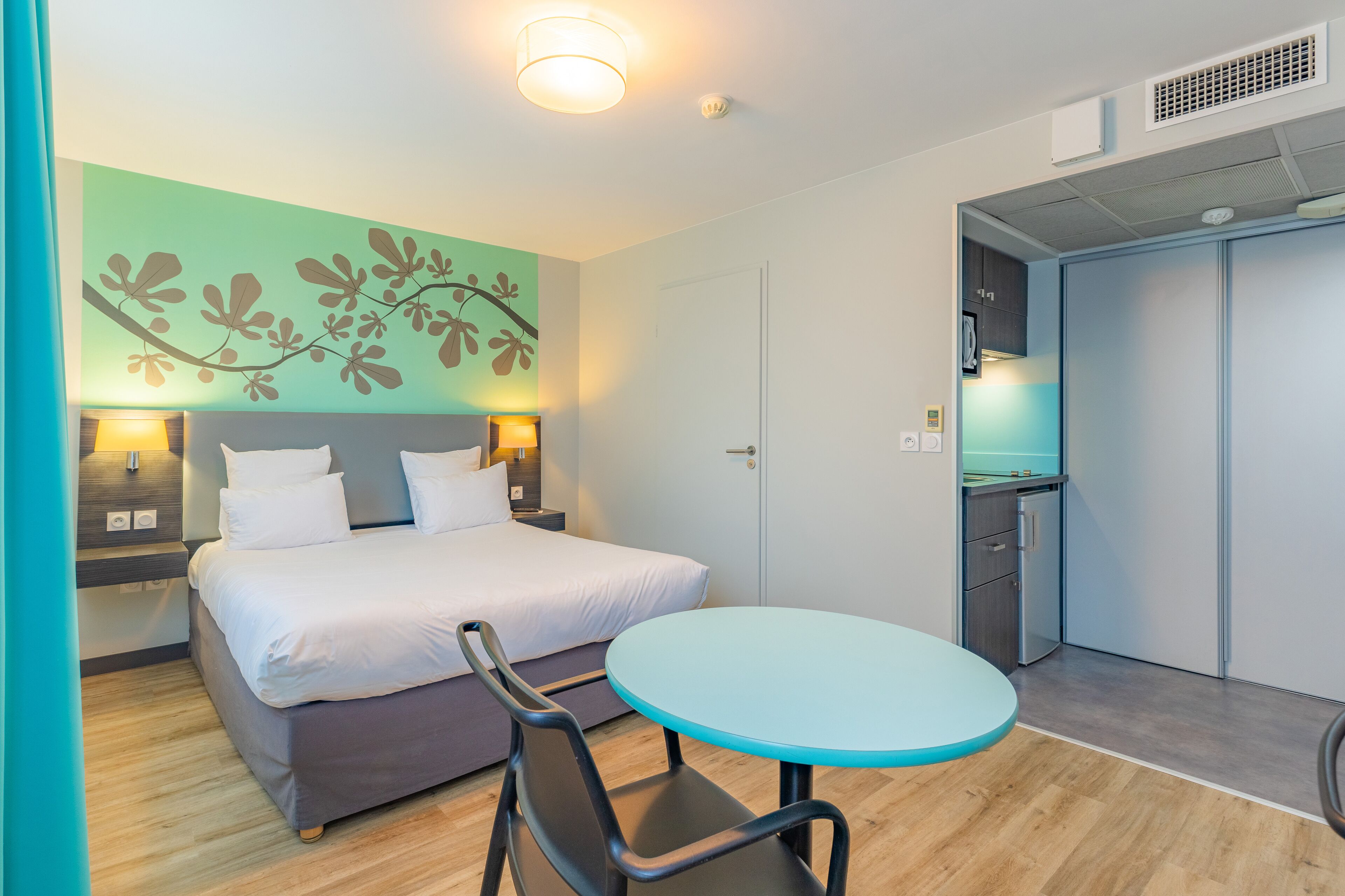 A hotel room with a bed, bedside tables, a round table, and a kitchenette.
