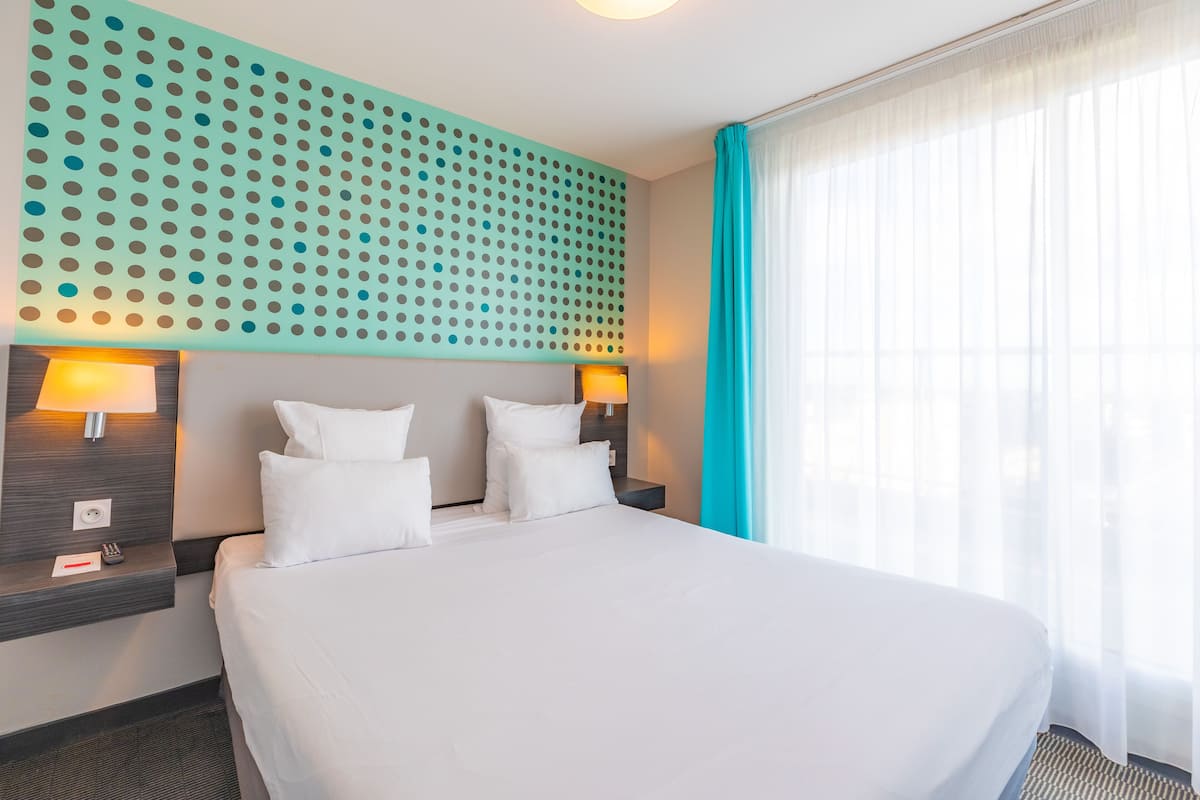 A hotel room with a large bed, two bedside tables with lamps, a wall with a dot pattern, and a window with curtains.