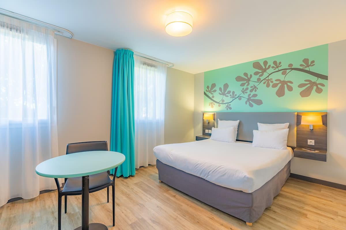 A hotel room with a bed, a table, and a chair. There is a window with curtains, a wall with a floral design, and a nightstand with a lamp.