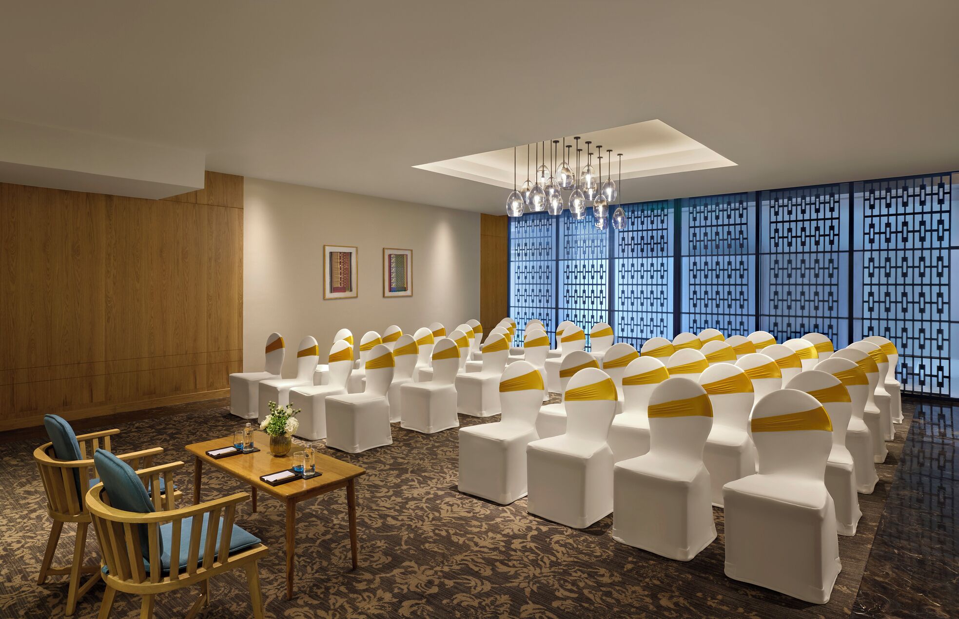A conference room with rows of white chairs with yellow sashes, a wooden table with a vase and glasses, and a patterned carpet.