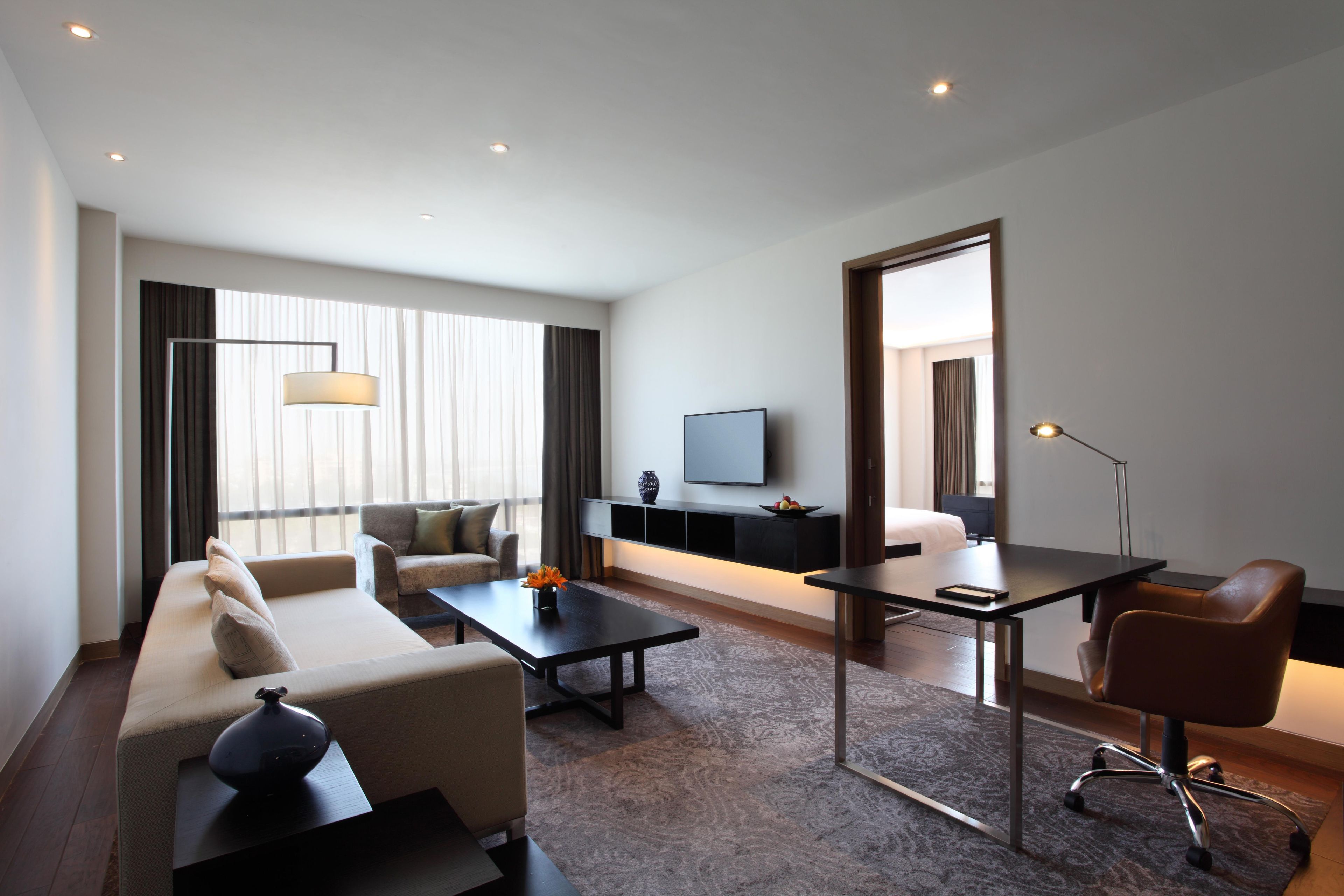 Una habitación de hotel moderna con sofá, una mesa de centro, escritorio, silla, televisor y un ventanal con cortinas.