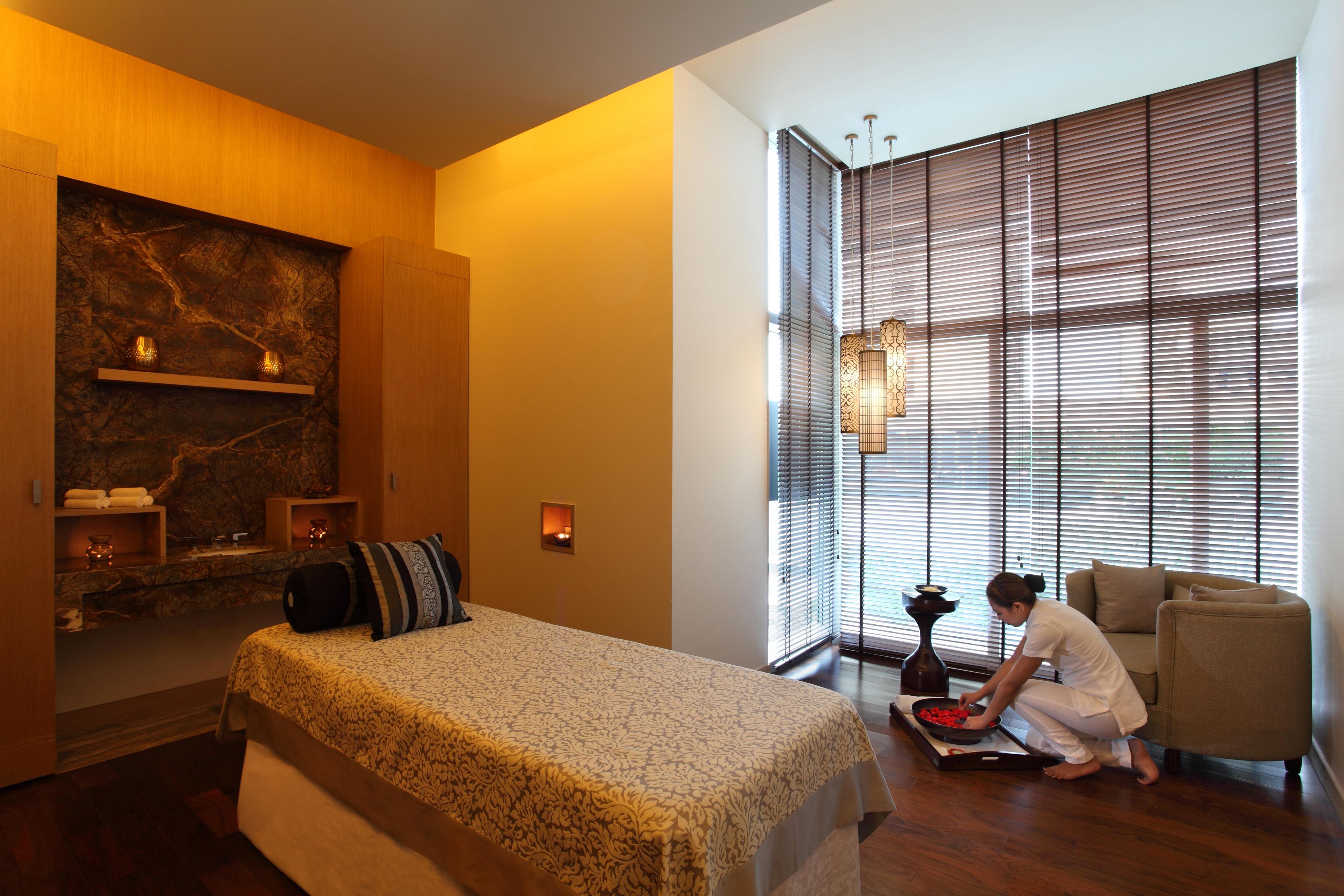 A hotel room with a bed, a massage table, a massage therapist, a window with blinds, and a stone wall.