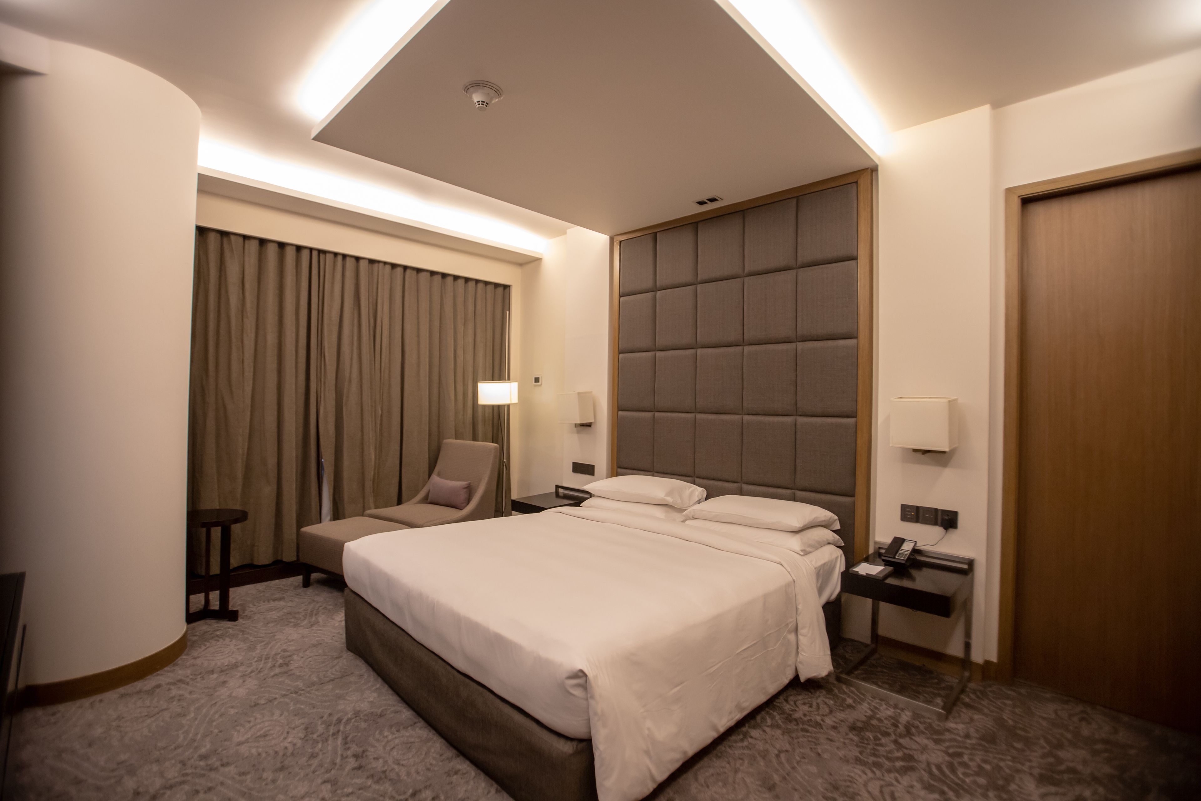 Habitación de hotel con una cama grande, una mesita de noche con lámpara, una silla y un ventanal con cortinas.