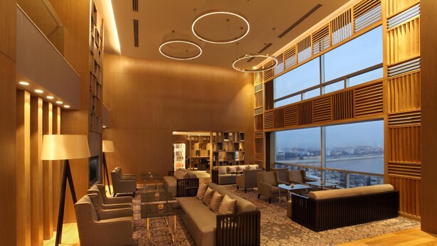 A modern lounge with sofas, armchairs, and a coffee table, large windows offering a view of the city, and a wooden accent wall.
