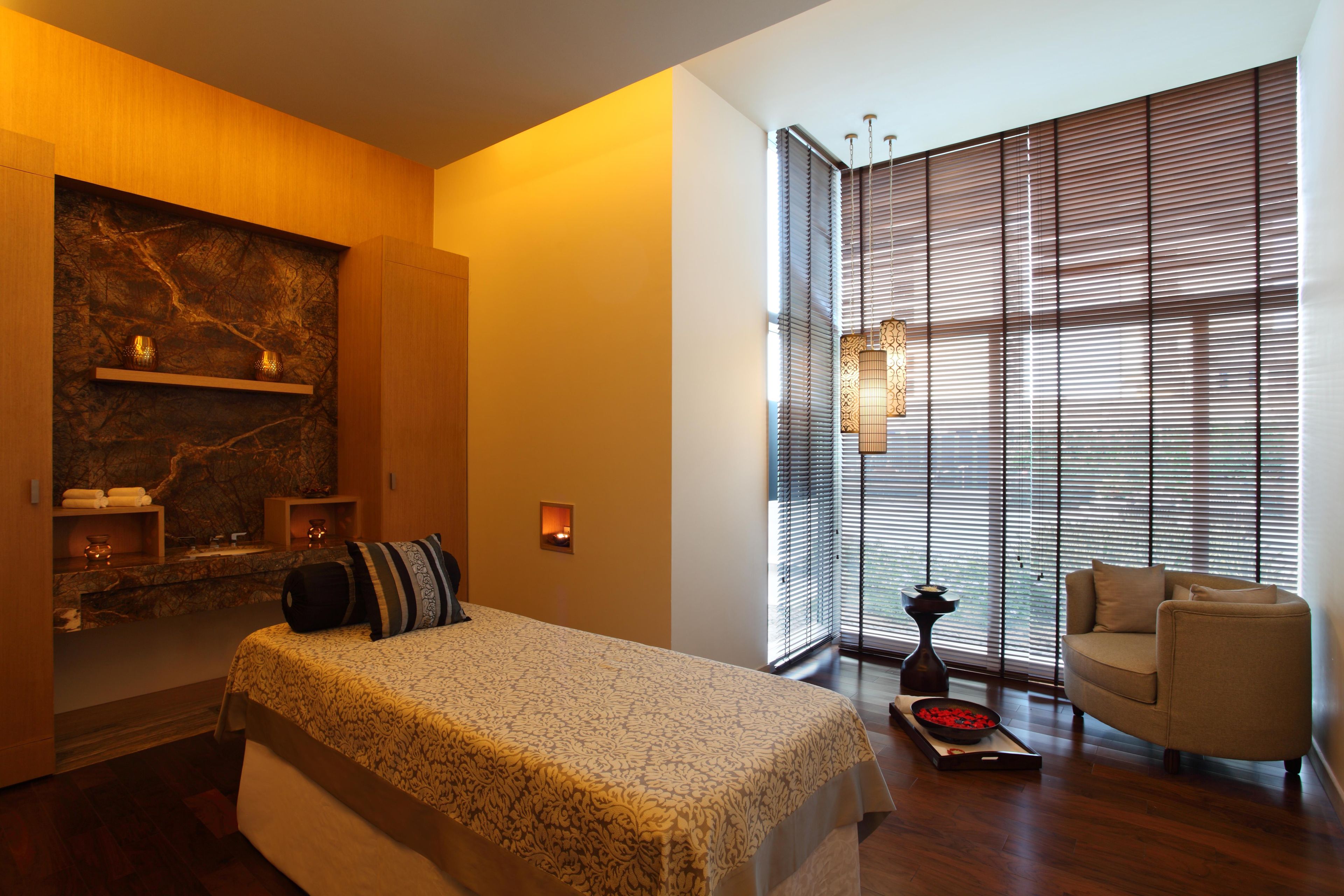 A modern bedroom with a bed, a seating area, a fireplace, and large windows with blinds.