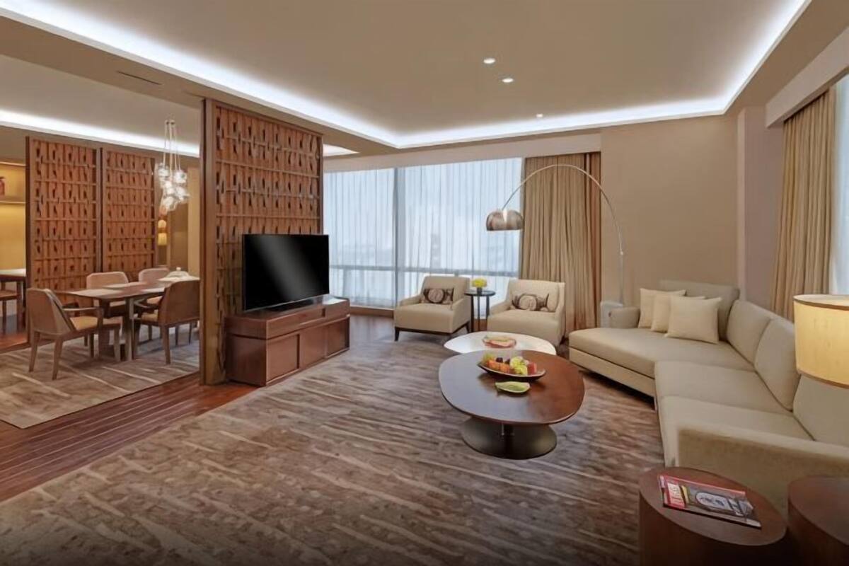 A modern hotel room with a flat-screen TV, a sofa, a dining area with chairs, and a large window.