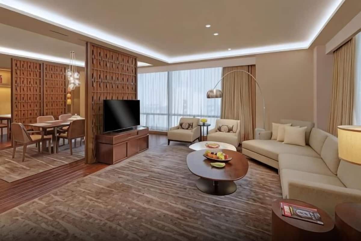 A modern hotel room with a flat-screen TV, a sofa, a dining area with chairs, and a large window.