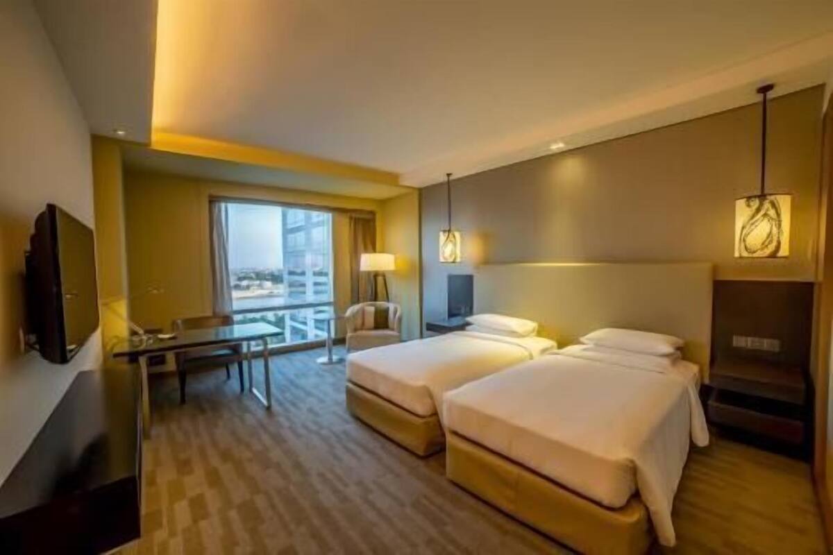 A hotel room with a bed, bedside table, lamp, TV, and a view of the city.