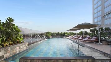 Una piscina con fuente acuática, rodeada de sillas de descanso y sombrillas, con un edificio al fondo.