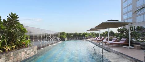 A swimming pool with a water feature, surrounded by lounge chairs and umbrellas, with a building in the background.