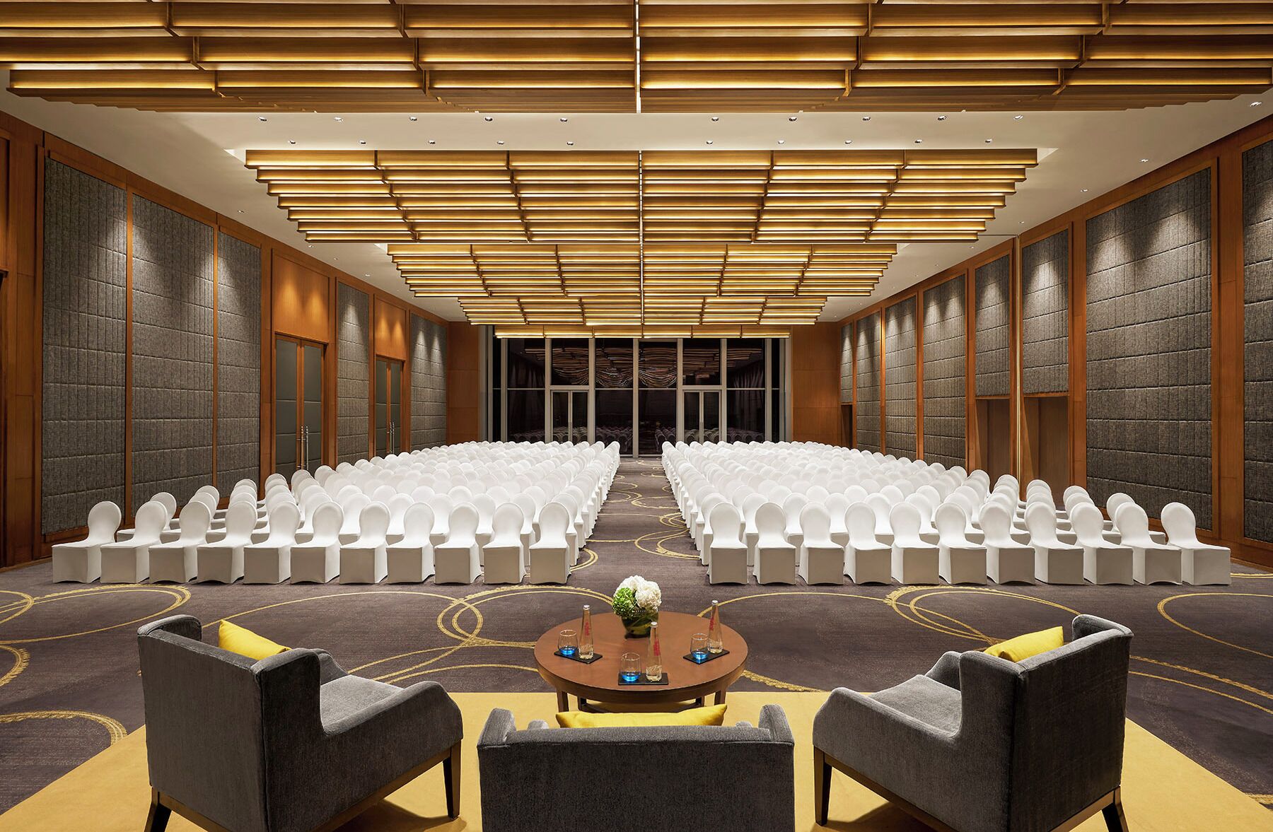 A conference room with rows of white chairs, a central table with a vase and glasses, and a modern ceiling design.