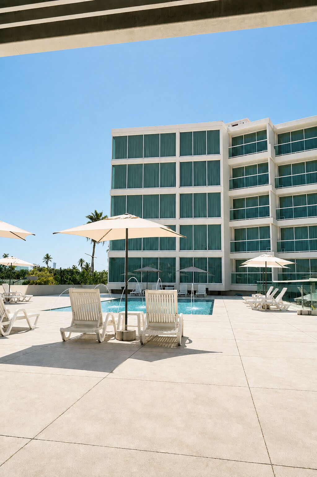 Un área de piscina con sillones de descanso y sombrillas, un edificio moderno de varios pisos con amplias ventanas, y un cielo azul despejado.