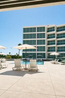 Un área de piscina con sillones de descanso y sombrillas, un edificio moderno de varios pisos con amplias ventanas, y un cielo azul despejado.