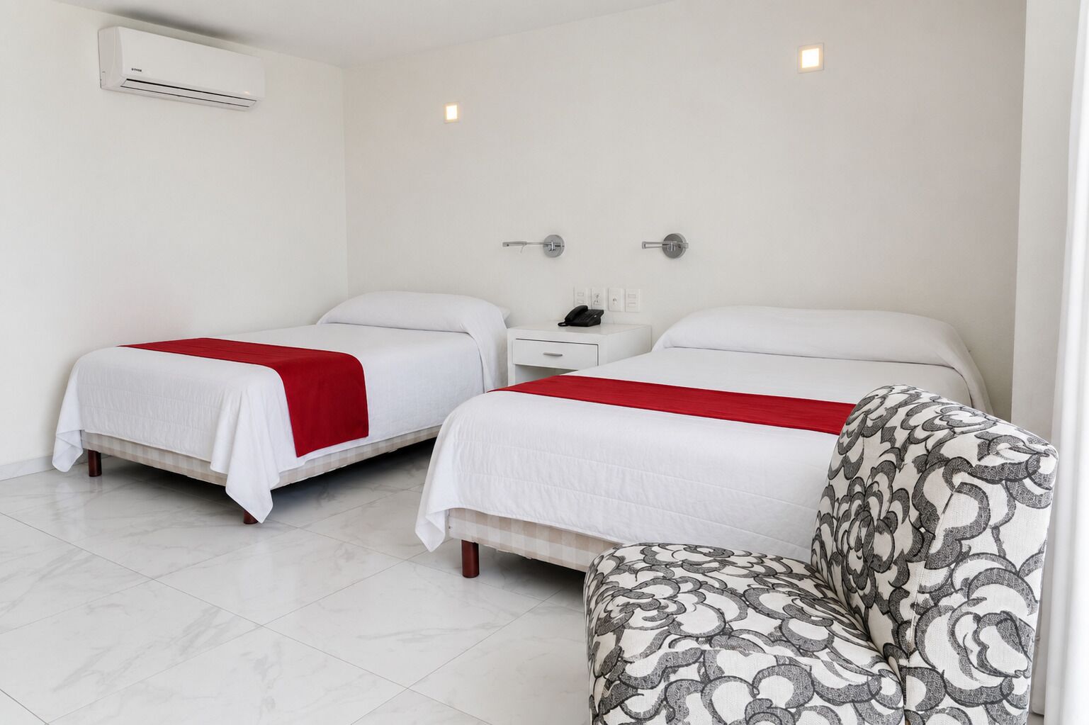 A hotel room with two beds, a bedside table with a phone, and a patterned armchair.