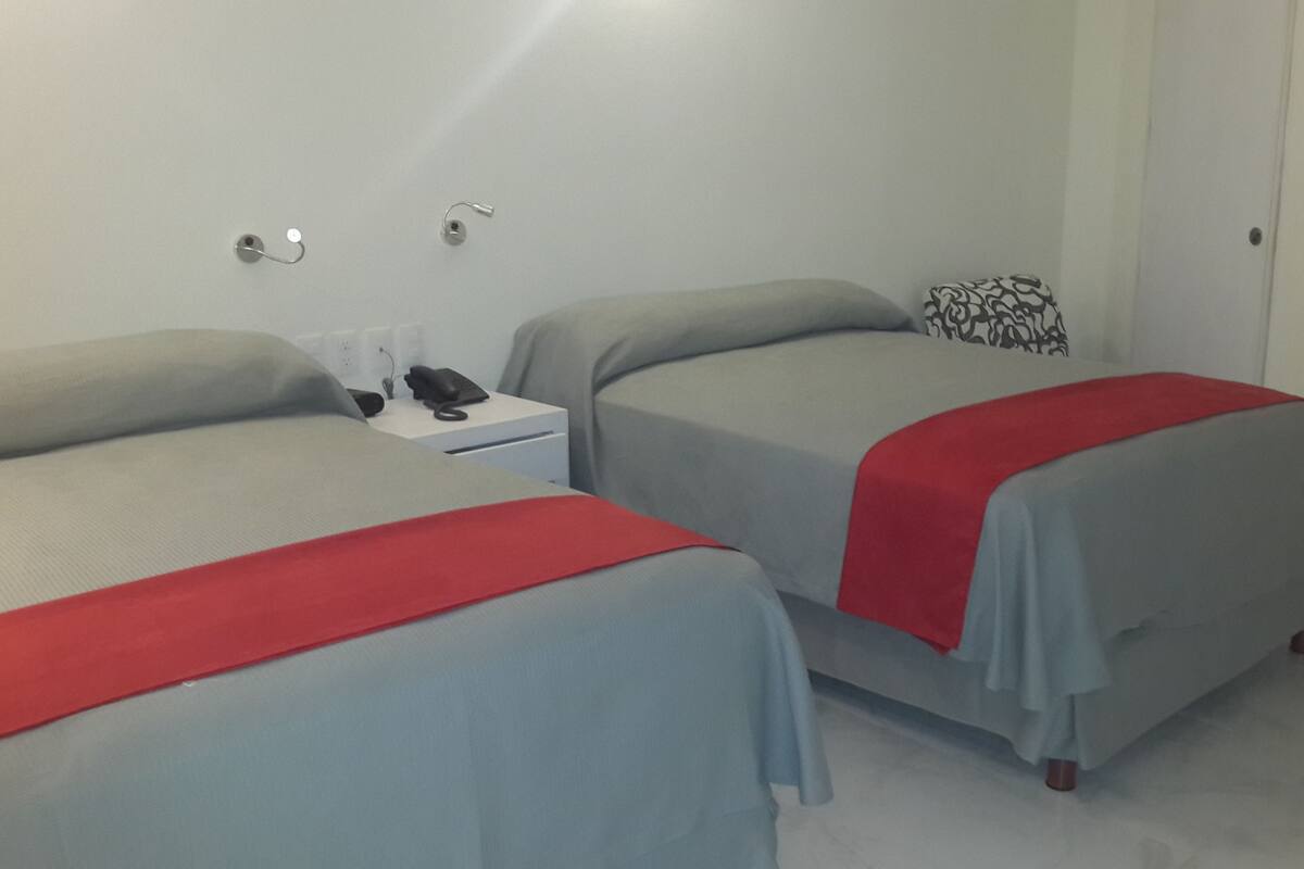 A hotel room with two beds, each with a red blanket, a bedside table with a phone and a lamp, and a wall socket.