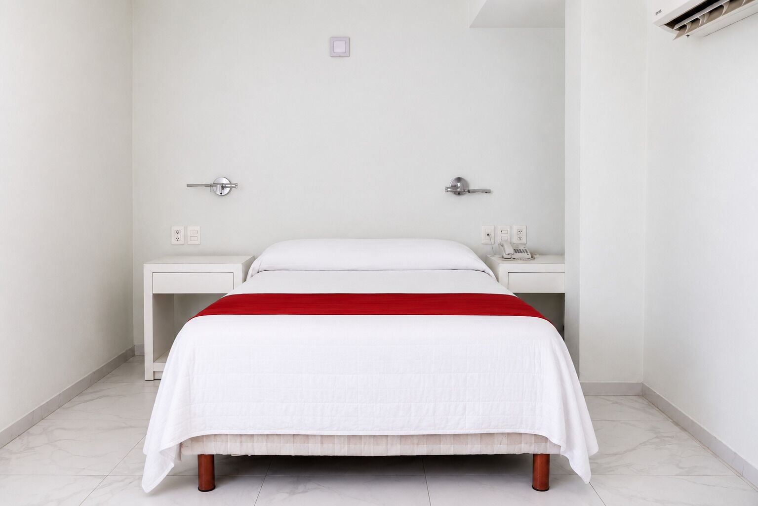 Habitación de hotel con cama, dos mesitas de noche y un detalle rojo en la colcha.