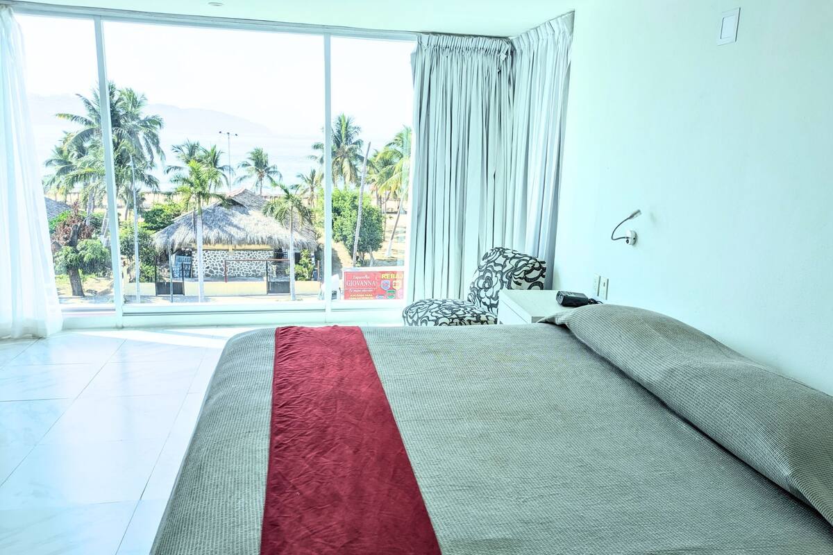 A hotel room with a large bed, a view of palm trees and a beach, and a thatched-roof building in the background.