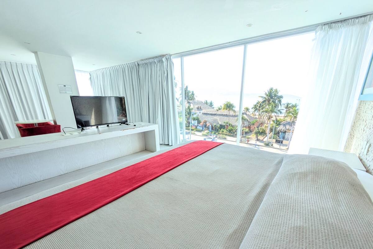 A modern hotel room with a large bed, a flat-screen TV on a white console, and a view of palm trees and buildings through large windows.