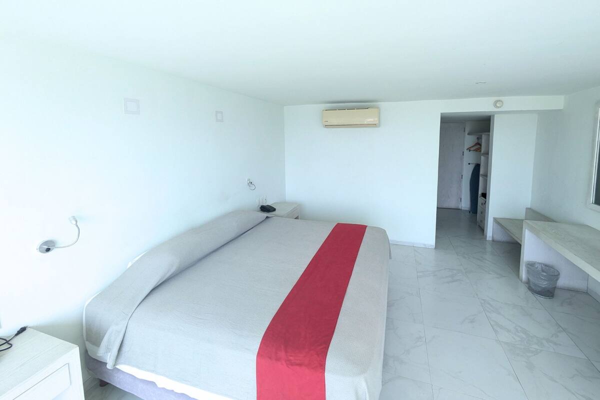 A hotel room with a bed, a desk, a chair, an air conditioning unit, and a bathroom visible through an open door.