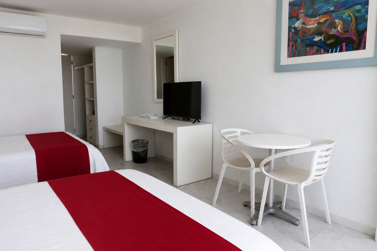 Habitación de hotel con dos camas, un escritorio con televisor, una pequeña mesa con sillas y un cuadro en la pared.