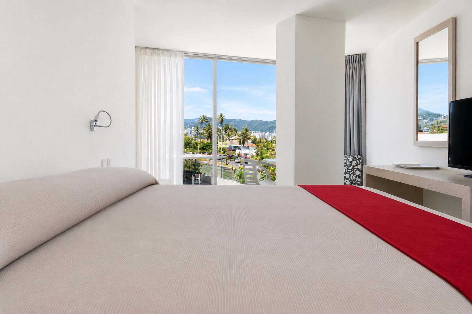 Habitación de hotel con una cama grande, televisión, escritorio y balcón con vista a la ciudad.