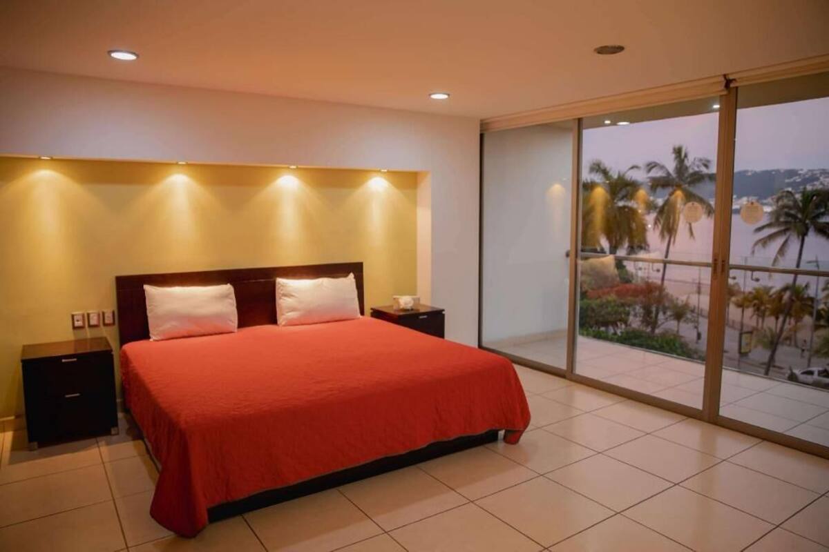 A bedroom with a large bed, a nightstand, a bedside table, and a view of palm trees through a sliding glass door.