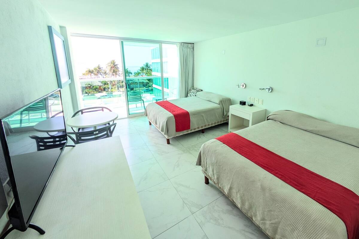 A hotel room with two beds, a desk, and a balcony with a view of the pool and palm trees.