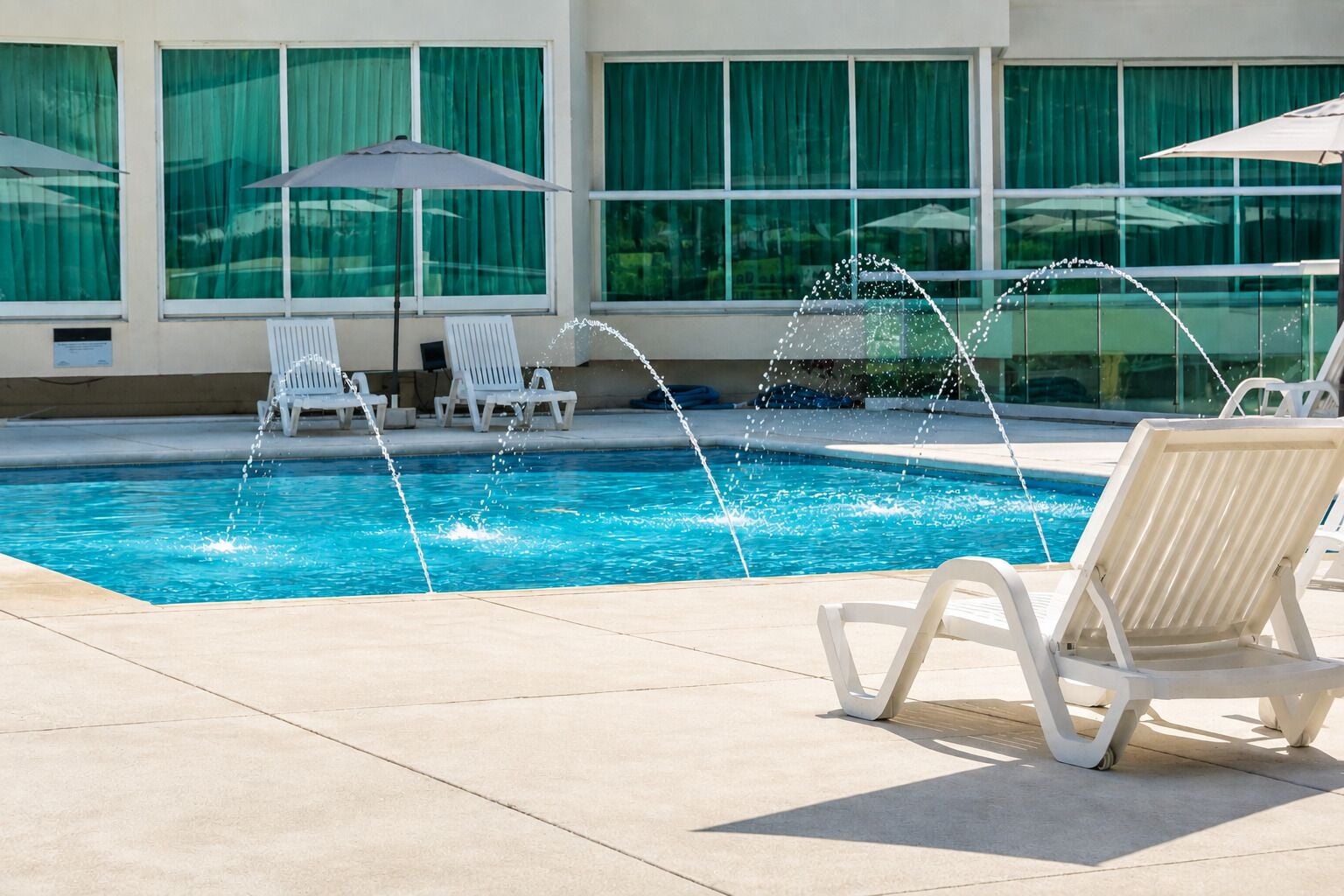 Una piscina con chorros de agua, sillas de descanso blancas y sombrillas.
