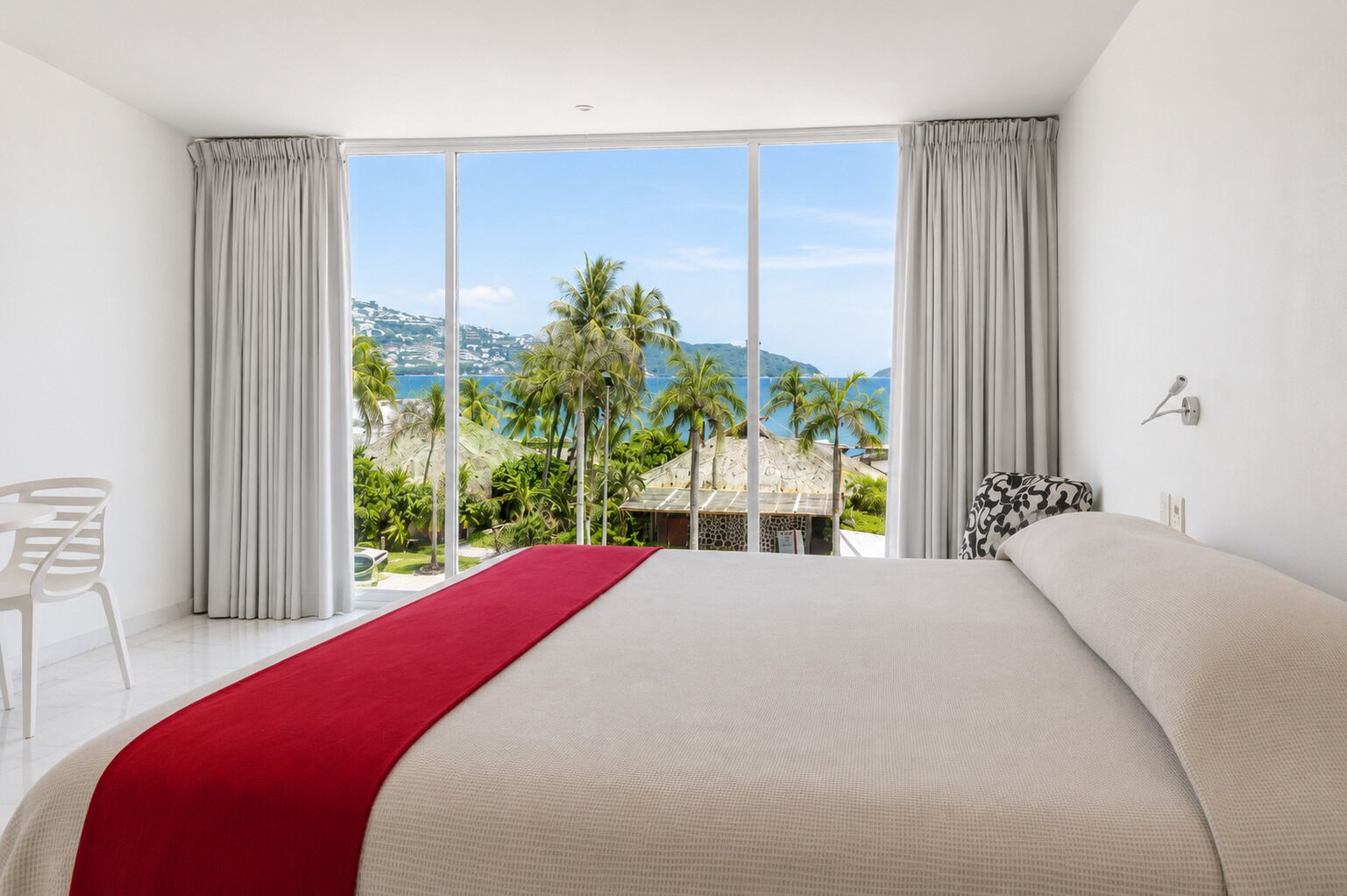 Habitación de hotel con una cama grande, un toque de color rojo, vistas a palmeras y montañas, y una silla blanca.