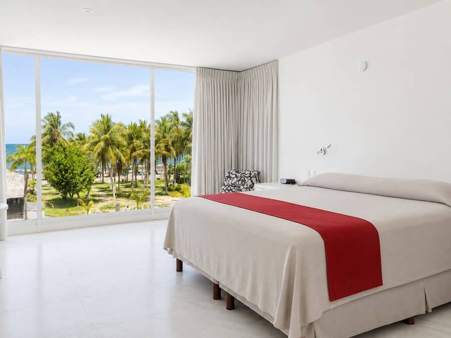 Una habitación de hotel con una cama grande, vistas a palmeras y al océano, un escritorio blanco y una ventana con cortinas.