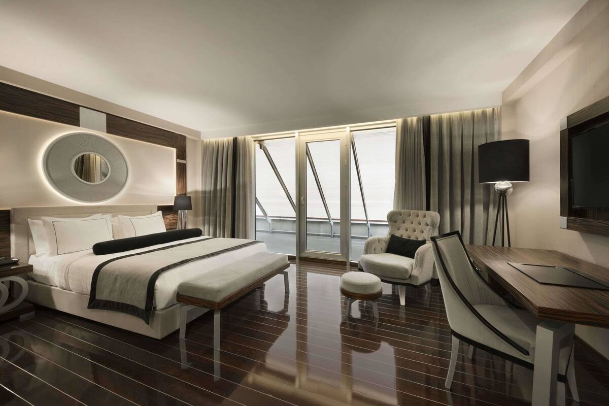 A modern hotel room with a large bed, a desk with a chair, a round mirror, and a television.