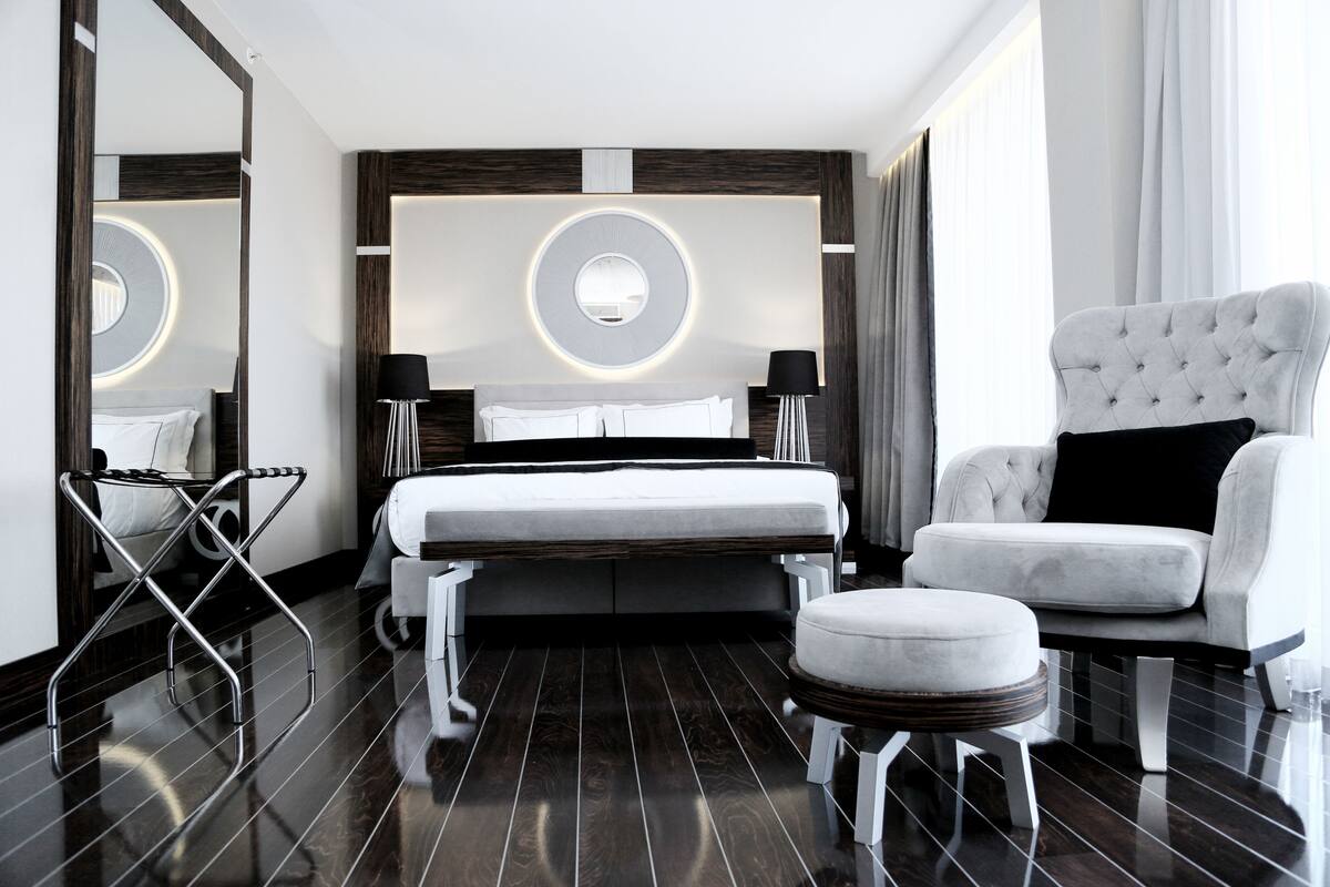 A modern hotel room with a large bed, a tufted armchair, a round mirror, and a side table.