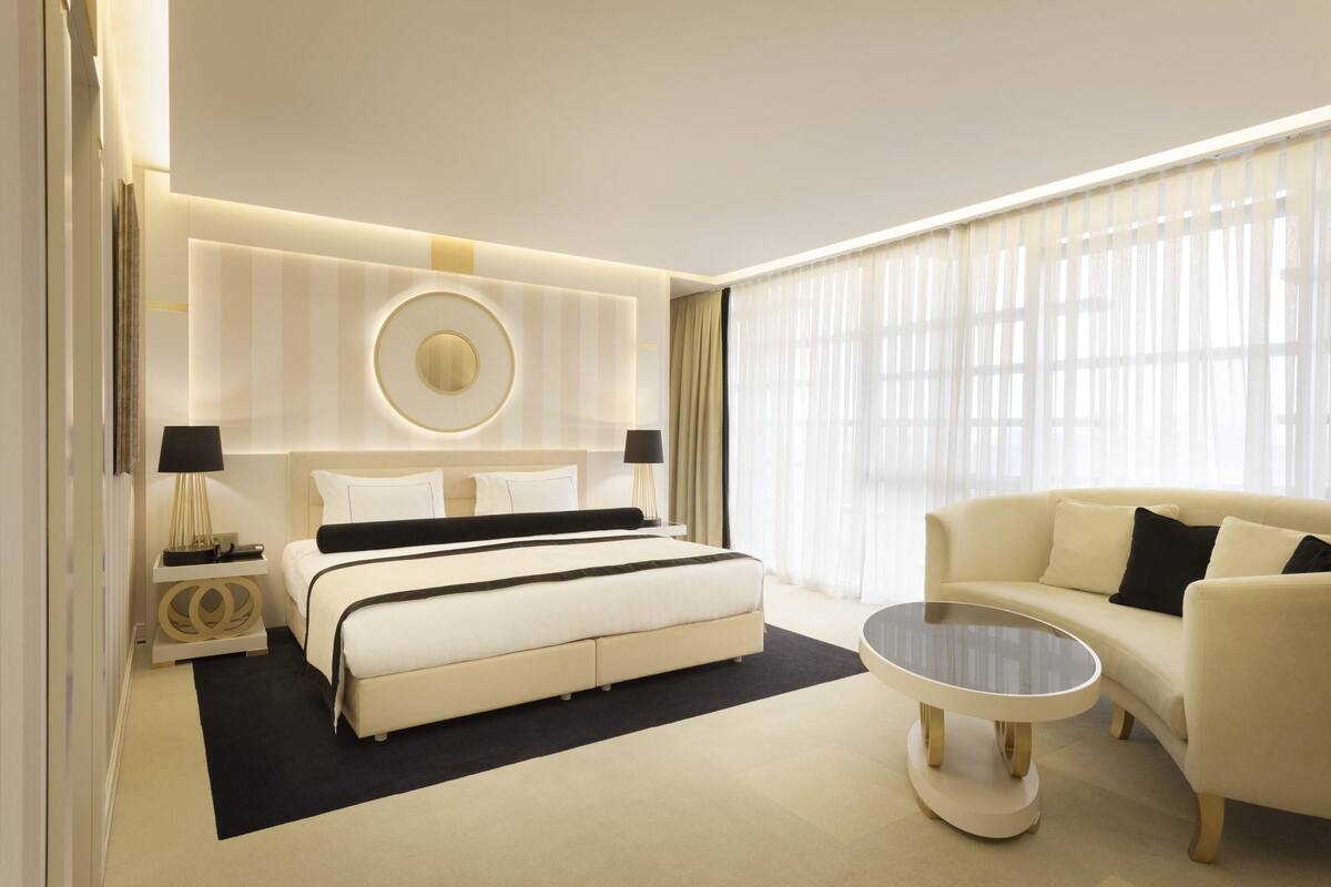 A modern hotel room with a large bed, a sofa, a round coffee table, and a large window with sheer curtains.