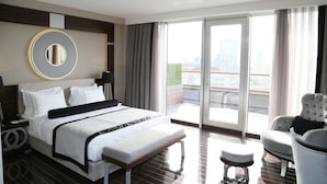 Una habitación de hotel moderna con una cama grande, un espejo redondo, una zona de estar con una silla y un reposapiés, y un balcón con vistas a la ciudad.