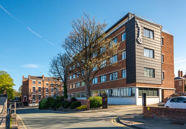 Un immeuble à plusieurs étages, doté d’un style architectural moderne, avec une façade combinant des éléments en brique et en métal, de grandes fenêtres et un arbre devant l’entrée.