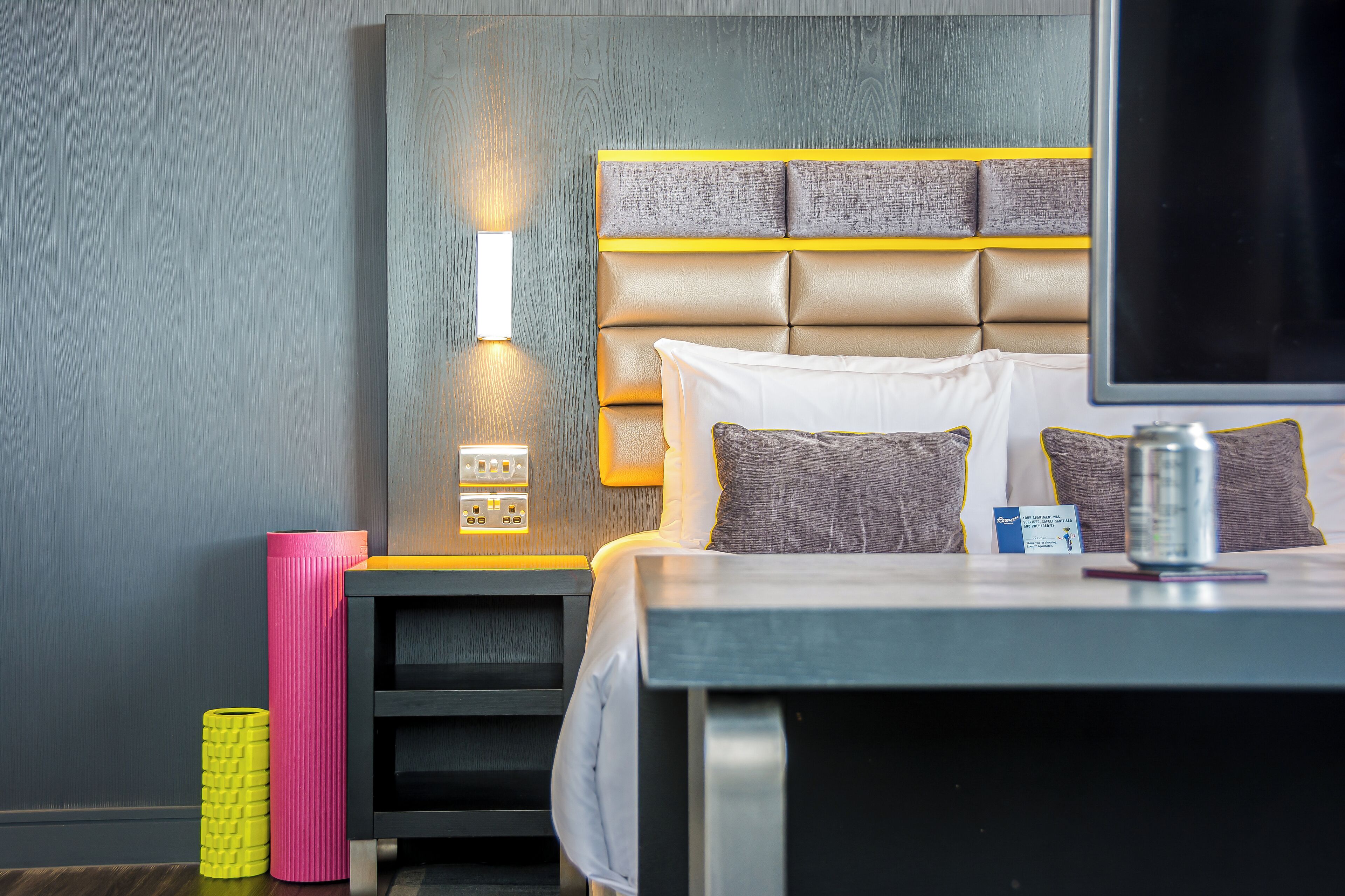 A modern hotel room with a large bed, a bedside table, a flat-screen TV, and a wall-mounted light fixture.