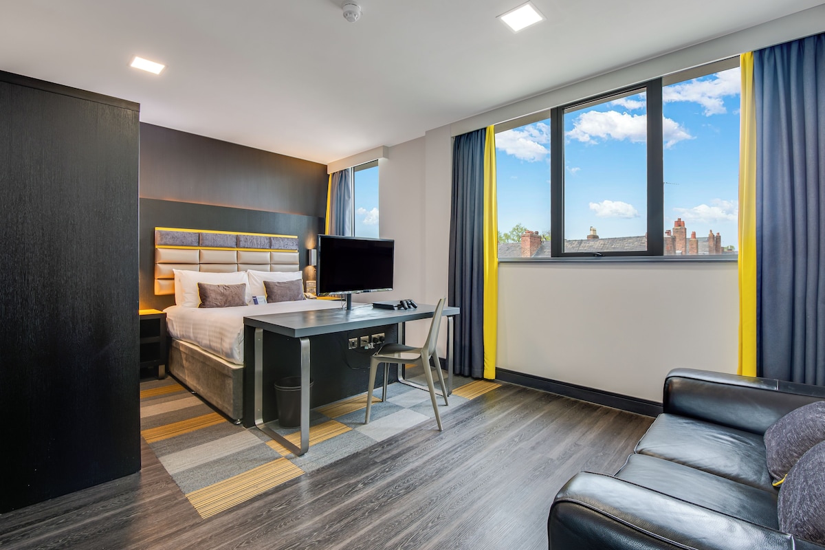 Ein modernes Hotelzimmer mit Bett, Schreibtisch, Stuhl und Blick auf die Skyline durch große Fenster.